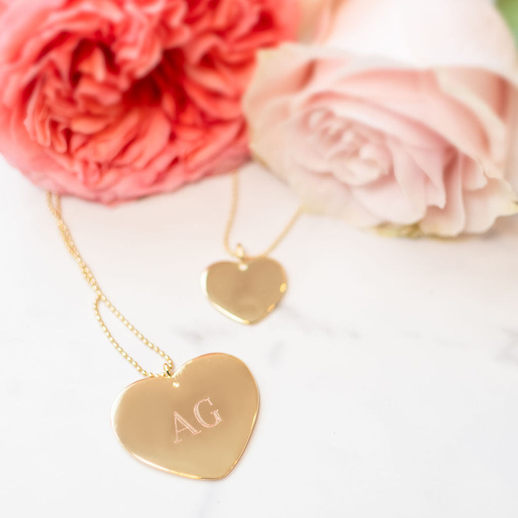Two gold-plated heart-shaped necklaces with one featuring a monogram 'AG' initial, displayed on a white surface with pink roses in the background.
