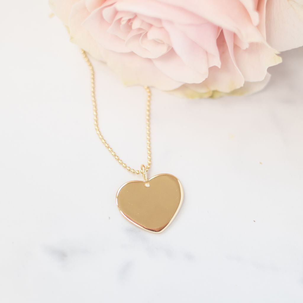 A gold-plated heart necklace on a 16-beaded chain, displayed against a white background with a pink flower to the side.